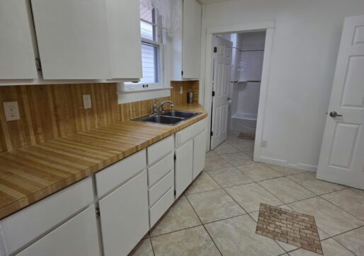 For Sale or Lease-222 Oak Ave, Eaton, CO 80615. A kitchen with white cabinets, wood-patterned countertops, a stainless steel sink, and tiled flooring. There's a window above the sink and an open doorway leading to another room with white walls.