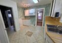 For Sale or Lease-222 Oak Ave, Eaton, CO 80615. A kitchen with green textured walls, white cabinets, tan tile flooring, a countertop with a stainless steel sink, and a microwave above a corner cabinet. An adjacent room with wood floors is visible through a doorway.