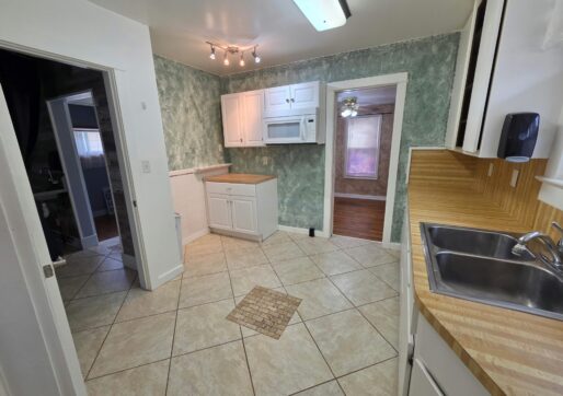 For Sale or Lease-222 Oak Ave, Eaton, CO 80615. A kitchen with green textured walls, white cabinets, tan tile flooring, a countertop with a stainless steel sink, and a microwave above a corner cabinet. An adjacent room with wood floors is visible through a doorway.
