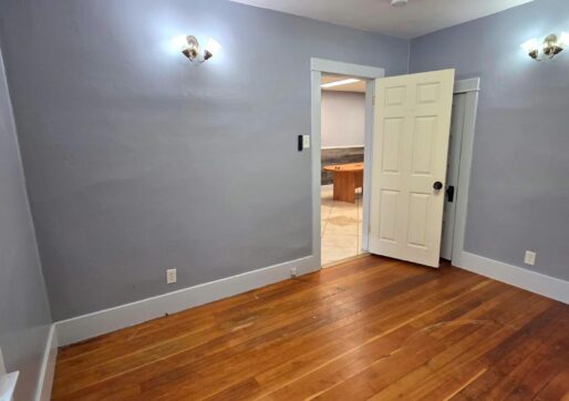 For Sale or Lease-222 Oak Ave, Eaton, CO 80615. An office with light gray walls, a wooden floor, two wall sconces, a ceiling light, and an open door leading to a tiled kitchen area.