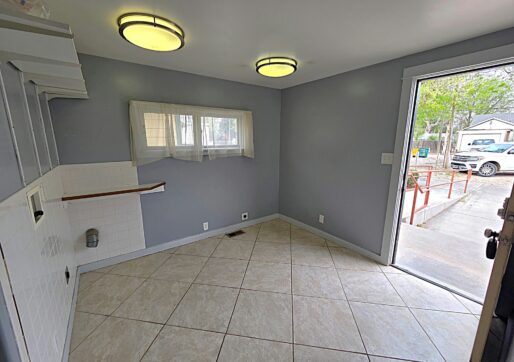 For Sale or Lease-222 Oak Ave, Eaton, CO 80615. A utility room with gray-blue walls, tiled floor, two ceiling lights, a window with sheer curtains, and an open door leading outside to a driveway with a parked car and trees visible.