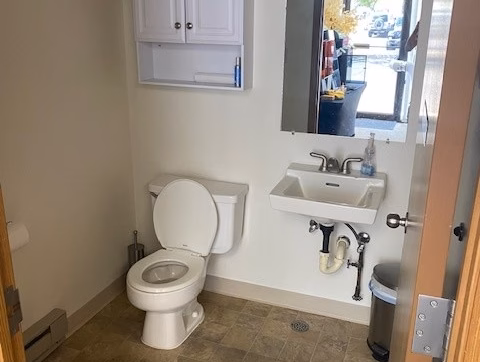 Office/Warehouse For Lease-Small bathroom with a toilet, wall-mounted white cabinet, sink, mirror, trash can, and a bouquet of yellow flowers on the cabinet. The door is open, showing a view outside through a window.