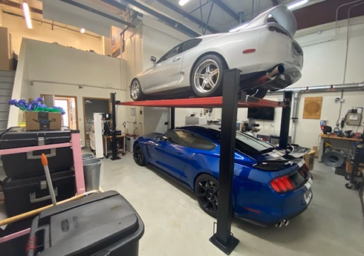 Office/Warehouse For Lease-A silver sports car is parked on a car lift above a blue sports car in a clean garage with storage bins and tools visible around the space.