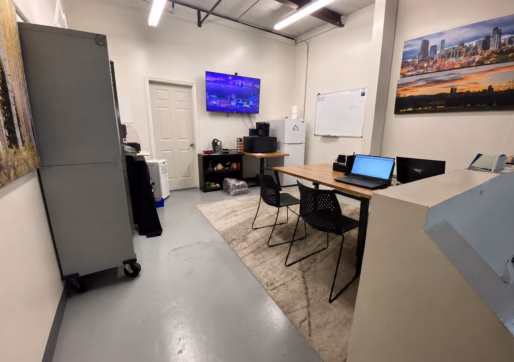 Retail Space For Lease-Modern office with a large table, three chairs, laptops, wall-mounted TV, whiteboard, storage cabinet, and cityscape artwork. Bright lighting and minimal decor create a clean, organized workspace—perfect for those seeking an Office/Warehouse For Lease.