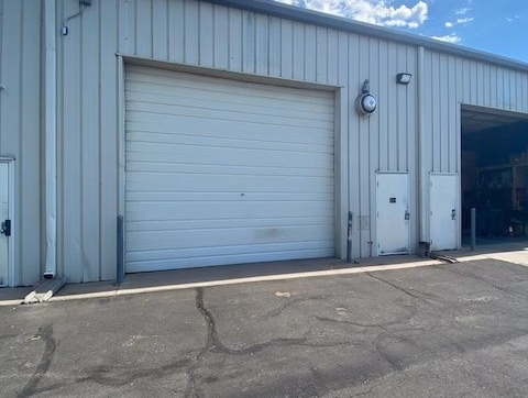 Office/Warehouse For Lease-A large closed white garage door on a metal building, with smaller doors and utility boxes nearby, under a partly cloudy blue sky. Asphalt pavement is in the foreground.