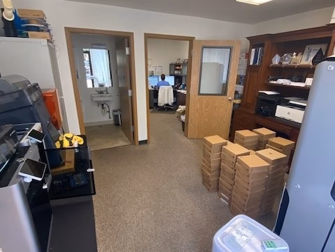 Office/Warehouse For Lease-Office with boxes stacked by an open door, desks with computers, printers on the left, a wooden cabinet on the right, and bright overhead lights. A person is visible working at a computer in the background.