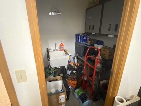 Office/Warehouse For Lease-Small utility closet with a sink, cleaning supplies, storage boxes, a red dolly, cabinets, and various tools and items stacked around the room.