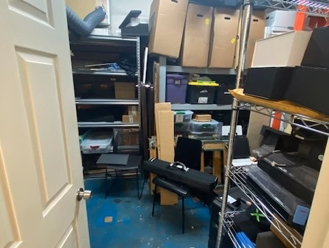 Office/Warehouse For Lease-A cluttered storage room with shelves, cardboard boxes, bins, and various items stacked and scattered on blue flooring. The door is open, revealing the crowded space.