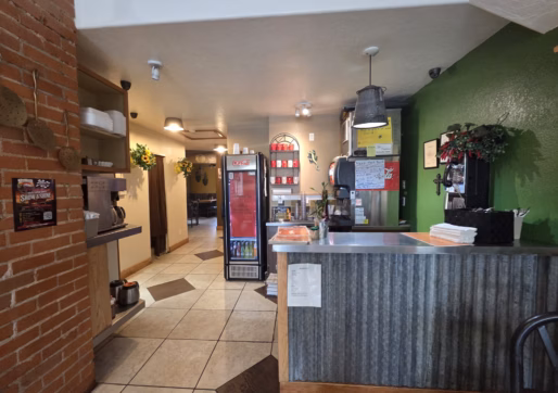 For Sale-226 Oak Ave, Eaton, CO-Counter at a restaurant with a green wall, stacked napkins, hanging menu, Coke fridge, and flowers. Kitchen area and tables are visible in the background on the left. Warm, cozy atmosphere.