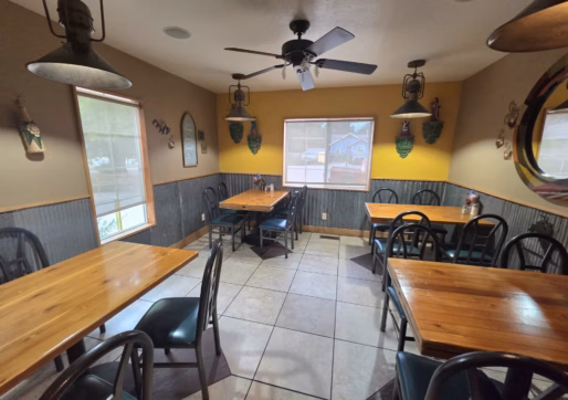 For Sale-226 Oak Ave, Eaton, CO - A small, cozy restaurant dining area with wooden tables, black chairs, wall decorations, a ceiling fan, and two windows letting in natural light. The room has yellow and beige walls with tile flooring.