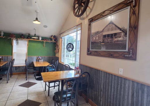 For Sale-226 Oak Ave, Eaton, CO - A cozy restaurant interior with tiled floors, wooden tables, green chairs, a wagon wheel decor, framed photo of a barn, and a large window letting in natural light.