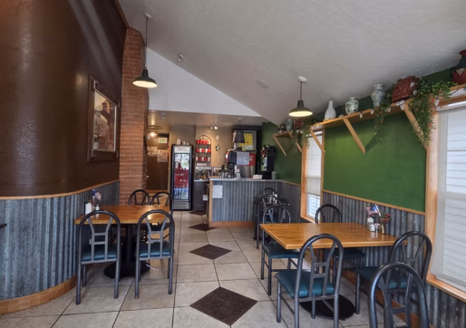 For Sale-226 Oak Ave, Eaton, CO - Cozy, small café with wooden tables, green and brown walls, tile floor, and metal chairs. Plants and decor line the shelves. A counter with a drink fridge is visible in the back. The space is well-lit by natural light.