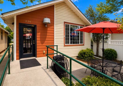 For Sale-226 Oak Ave, Eaton, CO - A small restaurant with a red and beige exterior, a glass door, and a ramp with green railings. Outdoor seating includes black tables, chairs, and a red umbrella. Trees and blue sky are visible.