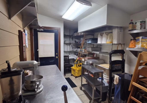 For Sale-226 Oak Ave, Eaton, CO - Commercial kitchen with stainless steel counters, shelves stocked with baking supplies, cleaning tools, a mop bucket, a blender, and a mixing bowl. Fluorescent lights illuminate the organized space.