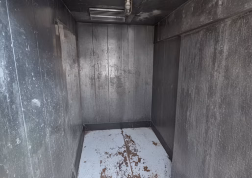 For Sale-226 Oak Ave, Eaton, CO - A small, empty metal room with gray walls and a white floor. A single light fixture is mounted on the ceiling. The space looks industrial.