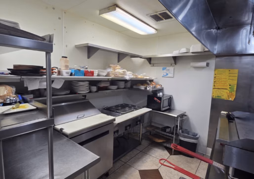 For Sale-226 Oak Ave, Eaton, CO - Small commercial kitchen with stainless steel counters, shelves holding dishes and food, a stove, fryer, prep area, and wall-mounted appliances under fluorescent lights. Floor has beige and brown tiles.