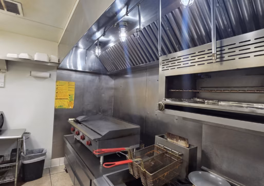 For Sale-226 Oak Ave, Eaton, CO - A commercial kitchen with a stove, fryer, griddle, pizza oven, metal backsplash, hanging utensils, and overhead lights. Plates are stacked, and a yellow sign is posted on the wall. The floor is tiled.