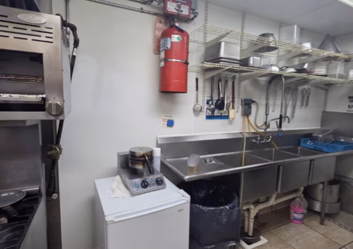 For Sale-226 Oak Ave, Eaton, CO - A commercial kitchen area with a sink, stainless steel shelves holding utensils and dishes, a fire extinguisher on the wall, a small fridge, and a trash can on tiled flooring.